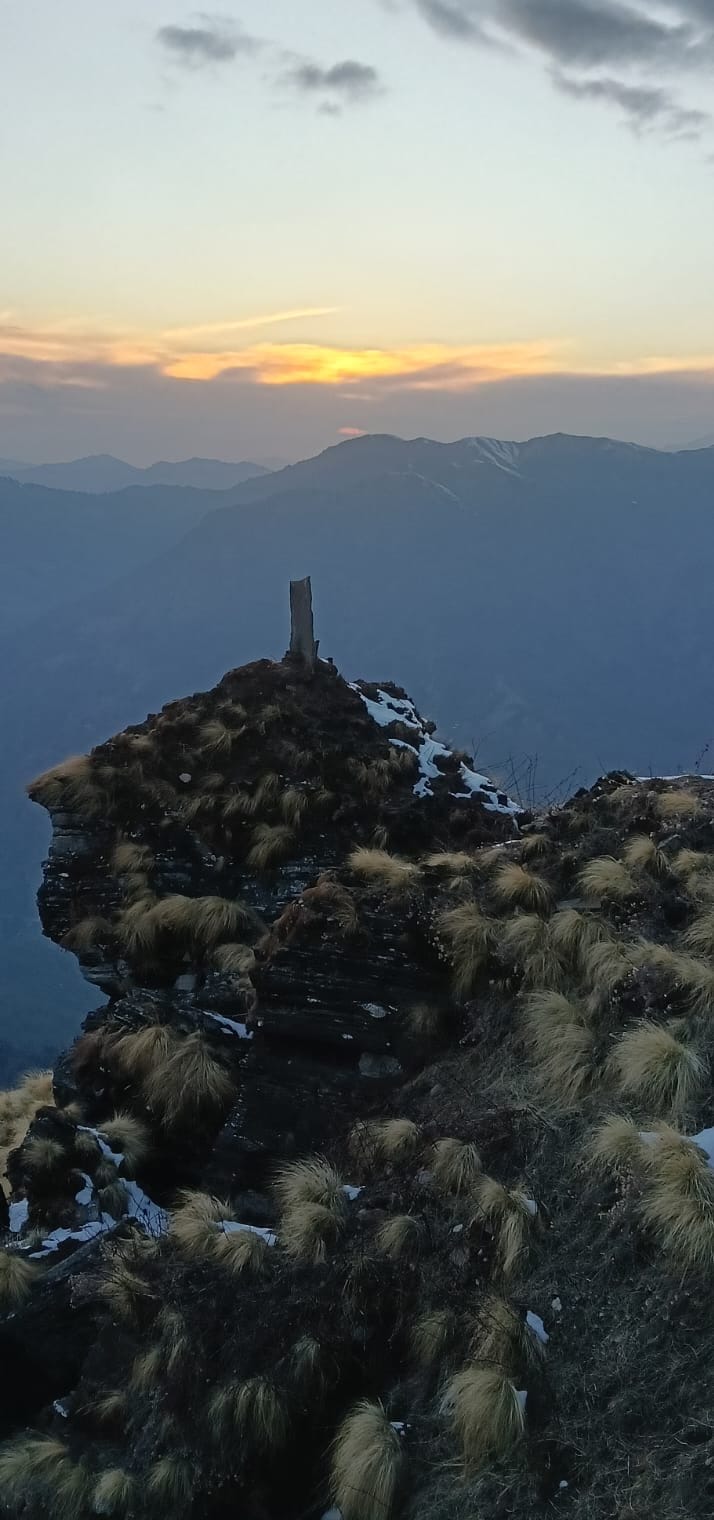 Sunset over layered Himalayan ridges seen from a rocky, grassy summit
